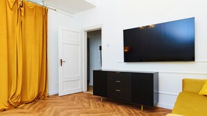 Una habitación con suelo de madera tipo espiga, un televisor negro en la pared, un sofá amarillo y una puerta blanca cerrada.