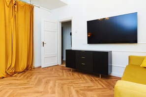 A room with a wooden herringbone floor, a black TV on the wall, a yellow sofa, and a closed white door. - Refined Central Sanctuary – Perfect Romantic Getaway by the River – 701 (Budapest)