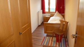 A bedroom with a wooden bed, a nightstand, a window with red curtains, and a radiator. - Schwendehof Vacation Home, Rental Unit 1 (Fontanella)