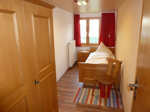 A bedroom with a wooden bed, a nightstand, a window with red curtains, and a radiator. - Schwendehof Vacation Home, Rental Unit 1 (Fontanella)