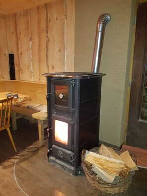 A wood-burning stove with a chimney, a wooden table, and a basket of firewood. - Schwendehof Vacation Home, Rental Unit 1 (Fontanella)
