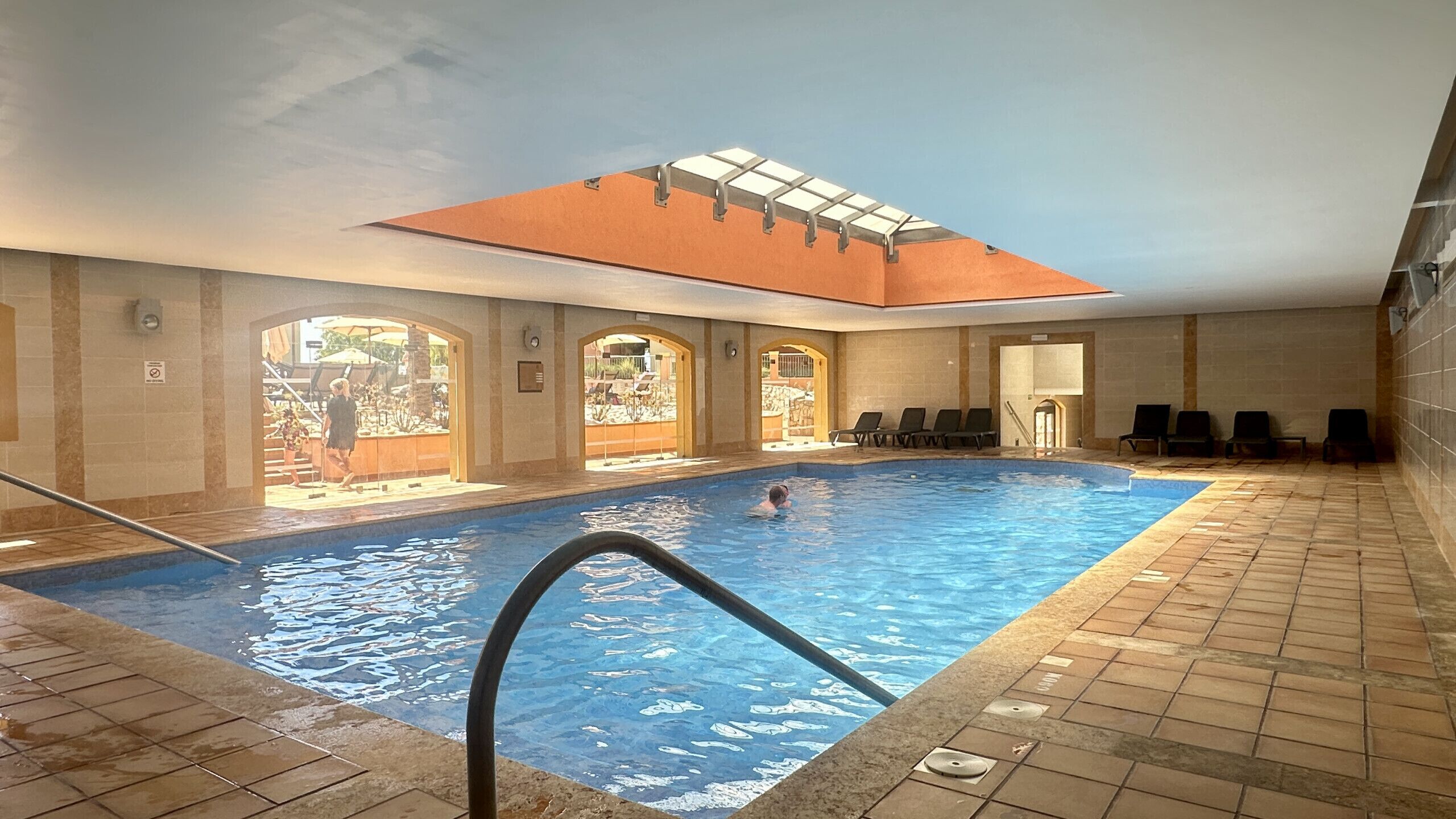 An indoor swimming pool with a skylight, tiled walls, and a sunlit area with chairs.