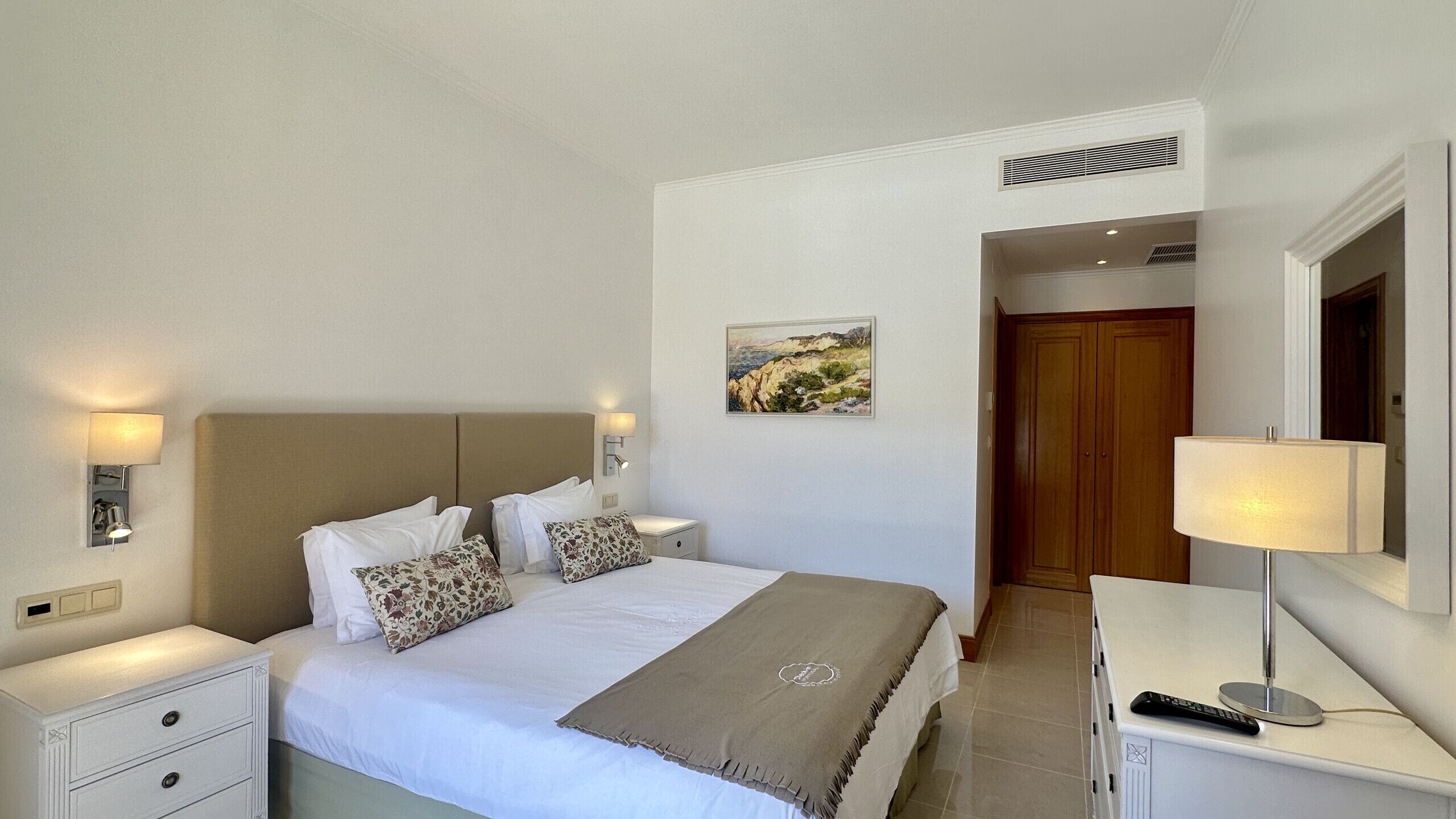 A hotel room with a bed, bedside tables, a nightstand, a lamp, a painting, and a wardrobe.