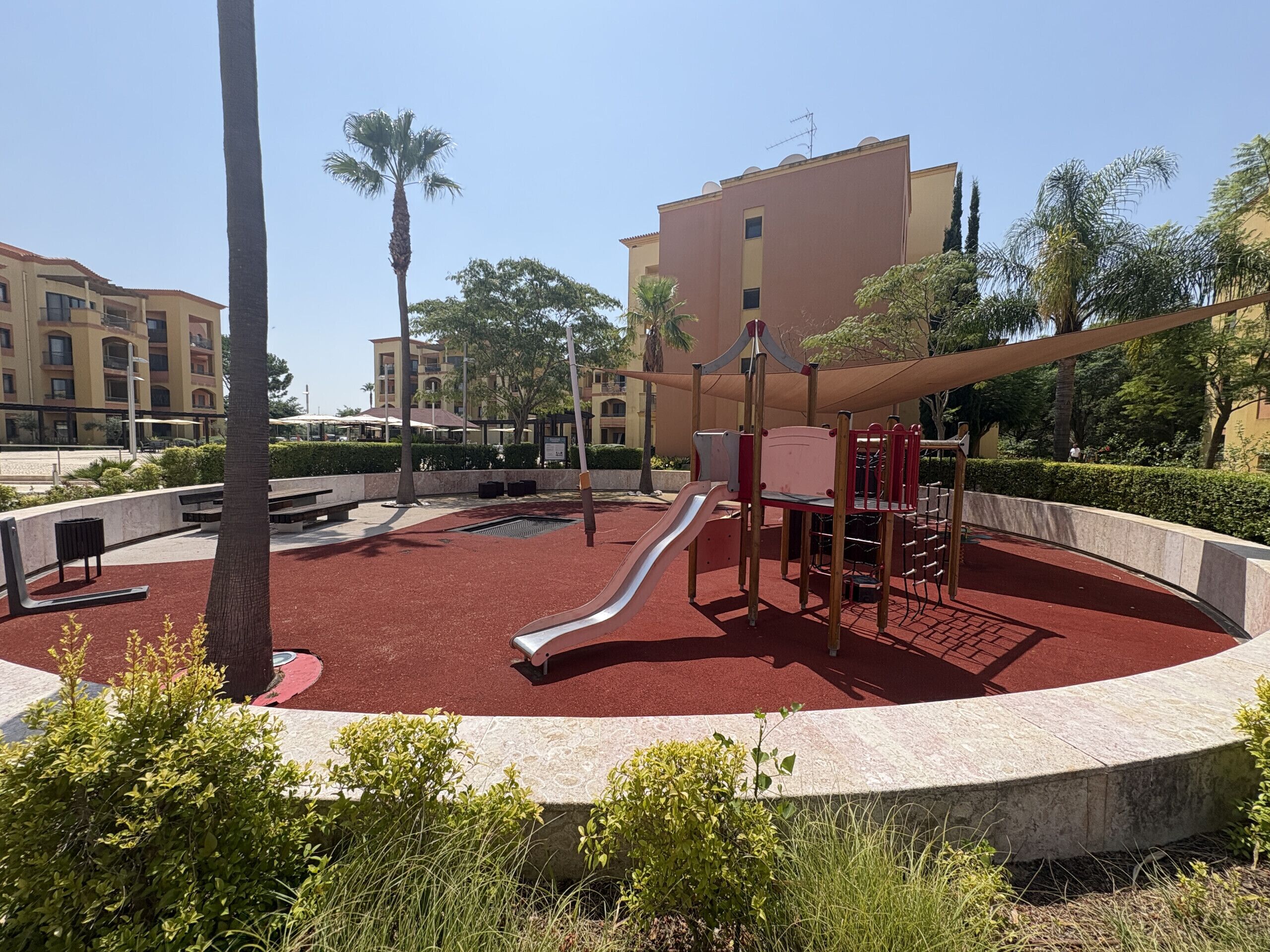 A playground with a slide and climbing structures, surrounded by a circular seating area and palm trees.