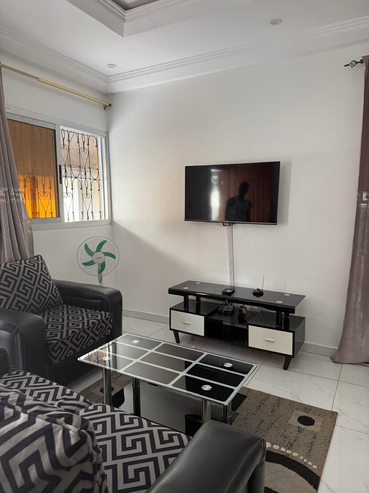 Un salon moderne avec un téléviseur à écran plat fixé au mur, un canapé en cuir noir, une table basse en verre et un tapis à motifs.