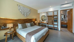 A modern bedroom with a wooden headboard, a large bed, a bedside table with a lamp, a wall-mounted macrame piece, a wooden desk with a chair, a mirror, and a wardrobe. - Villa Yuka (Ubud)