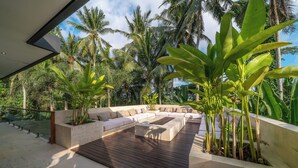 A modern outdoor seating area with a wooden deck, beige cushions, and a stone planter with tropical plants. - Villa Yuka (Ubud)