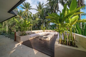 A modern outdoor seating area with a wooden deck, beige cushions, and a stone planter with tropical plants. - Villa Yuka (Ubud)