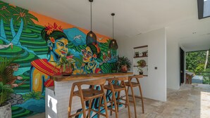 A modern kitchen with a vibrant mural, wooden bar stools, and potted plants. - Villa Yuka (Ubud)