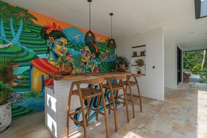 A modern kitchen with a vibrant mural, wooden bar stools, and potted plants. - Villa Yuka (Ubud)