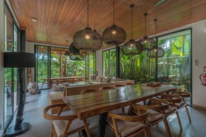 A spacious dining area with a long wooden table and chairs, surrounded by large windows offering a view of lush greenery. - Villa Yuka (Ubud)