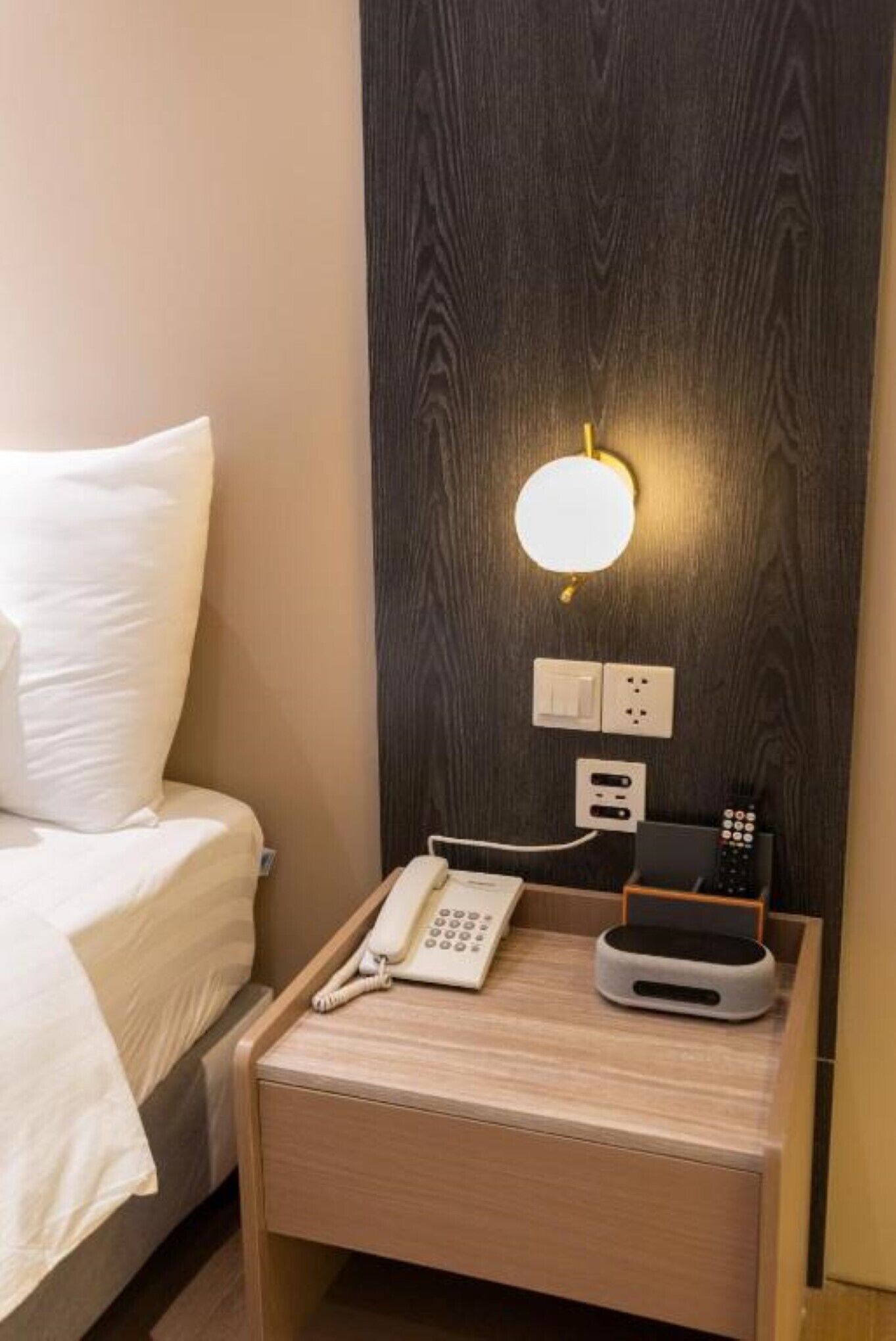 A hotel room with a bed, a bedside table with a phone, a remote control, and a wall-mounted light fixture.