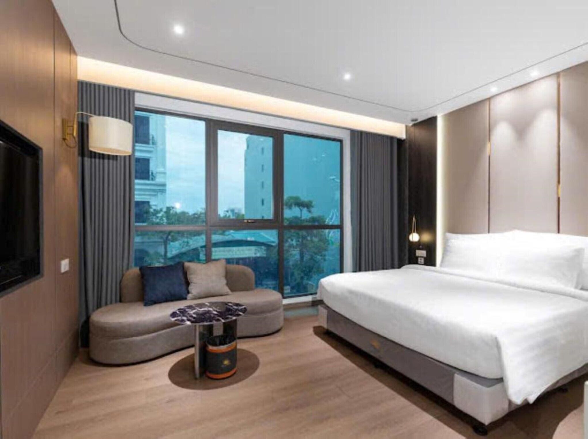 A modern hotel room with a large bed, a sofa, a small table, and a large window offering a city view.