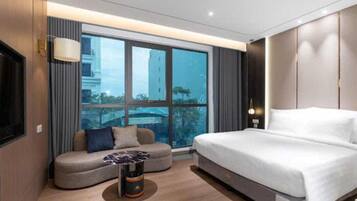 A modern hotel room with a large bed, a sofa, a small table, and a large window offering a city view.