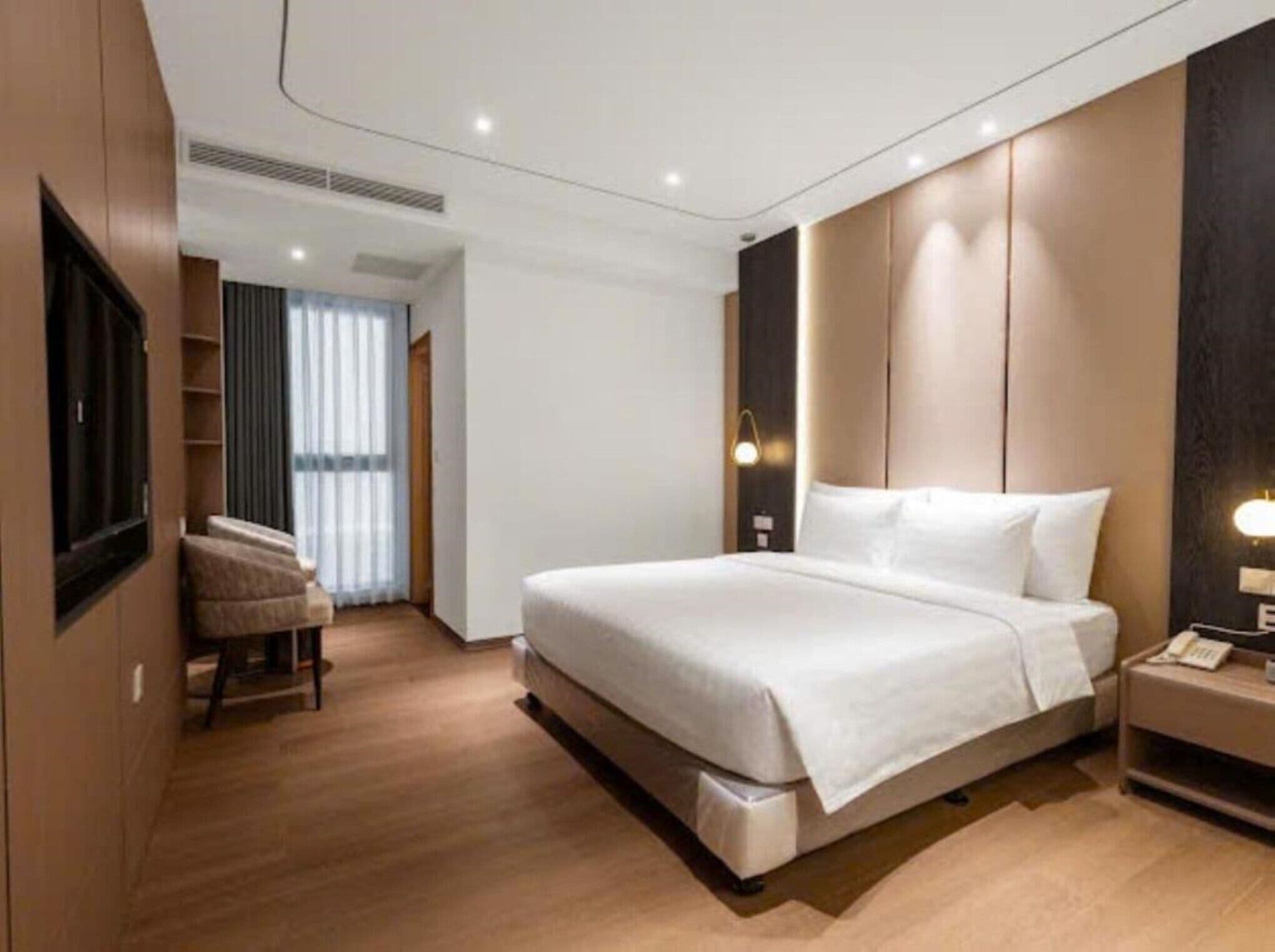 A modern hotel room with a large bed, a bedside table with a lamp, a chair, and a television mounted on the wall.