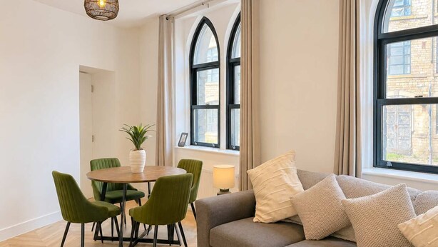 Un salon moderne avec une table à manger ronde, des chaises vertes, un canapé gris agrémenté de plusieurs coussins et de grandes fenêtres avec des rideaux.