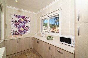 A kitchen with light-colored tiles, wooden cabinets, a microwave, and a window with a view of greenery. - Spacious Family Retreat with Two Houses & Private Pool (Javea)