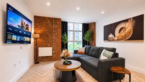 A modern living room with a flat-screen TV, a brick accent wall, a grey sofa, a round coffee table, and a large window. - Modern 1BR - Sleeps 4 - Free Parking - Manchester (Manchester)