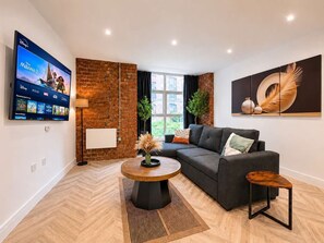 A modern living room with a flat-screen TV, a brick accent wall, a grey sofa, a round coffee table, and a large window. - Modern 1BR - Sleeps 4 - Free Parking - Manchester (Manchester)