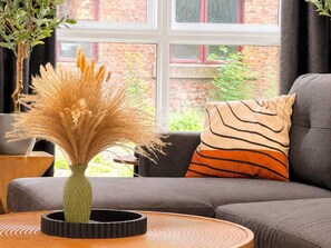 Ein Zimmer mit einer Couch, einem runden Tisch, einer Vase mit Trockenpflanzen und einem Fenster mit Blick auf ein Backsteingebäude.