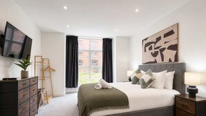 A modern hotel room with a large bed, a flat-screen TV, a desk with drawers, a chair, a window with curtains, and a painting on the wall. - Modern 1BR - Sleeps 4 - Free Parking - Manchester (Manchester)