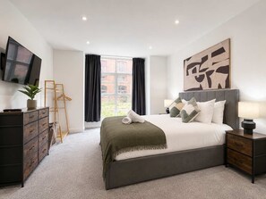 A modern hotel room with a large bed, a flat-screen TV, a desk with drawers, a chair, a window with curtains, and a painting on the wall. - Modern 1BR - Sleeps 4 - Free Parking - Manchester (Manchester)