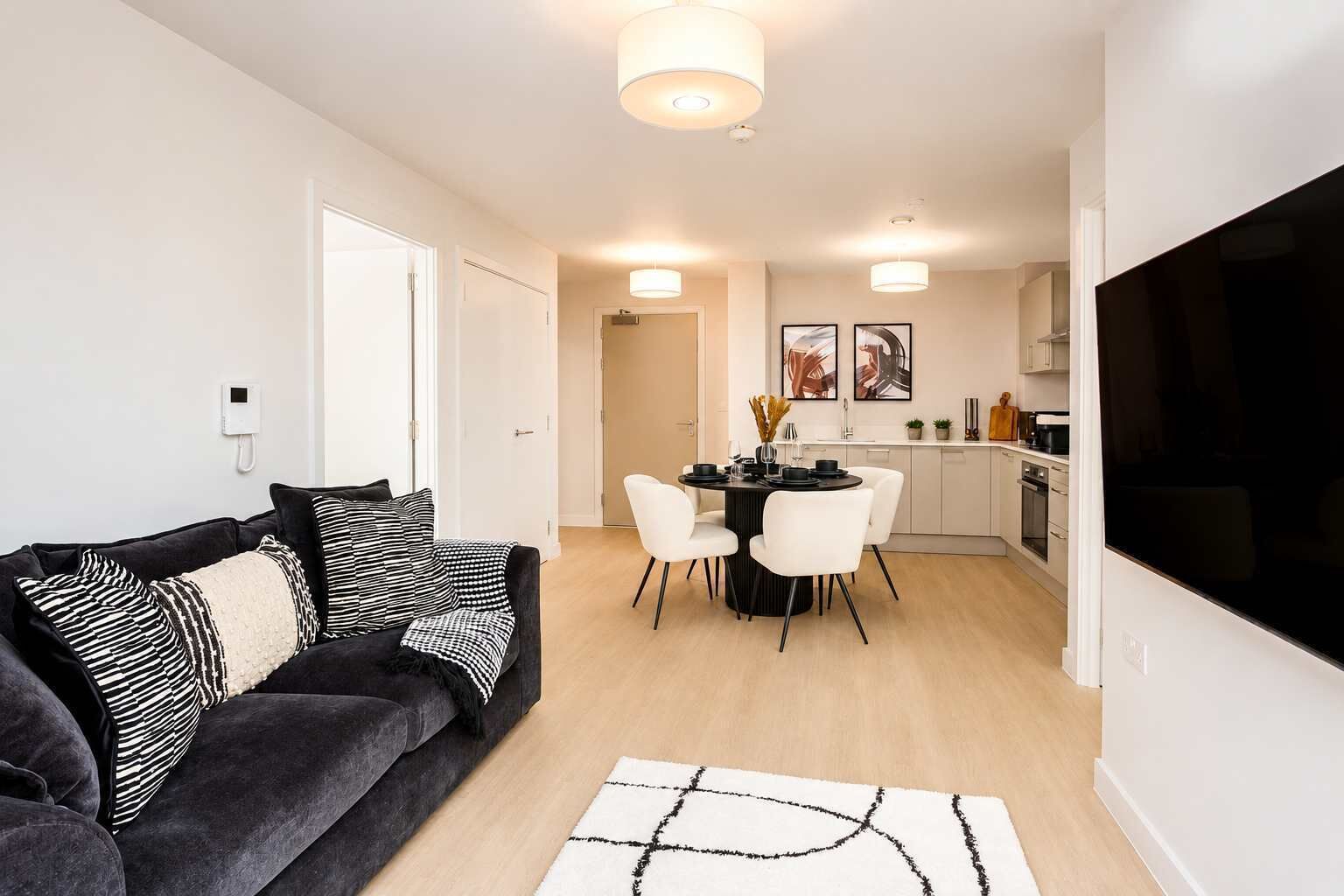 A modern living room with a dark grey sofa, a dining table with white chairs, and a kitchen area with white cabinets and a black TV.
