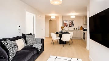 A modern living room with a dark grey sofa, a dining table with white chairs, and a kitchen area with white cabinets and a black TV.