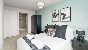 A modern bedroom with a bed, bedside tables, a nightstand, a wall-mounted lamp, and a door leading to a bathroom. - Luxury VIEWS!  10th  Floor  2 Bed Apartment (Manchester)