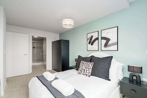 A modern bedroom with a bed, bedside tables, a nightstand, a wall-mounted lamp, and a door leading to a bathroom. - Luxury VIEWS!  10th  Floor  2 Bed Apartment (Manchester)