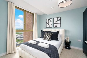 A modern bedroom with a large window, a bed with white bedding and black and white pillows, a black side table with a lamp, and two abstract art pieces on the wall. - Luxury VIEWS!  10th  Floor  2 Bed Apartment (Manchester)