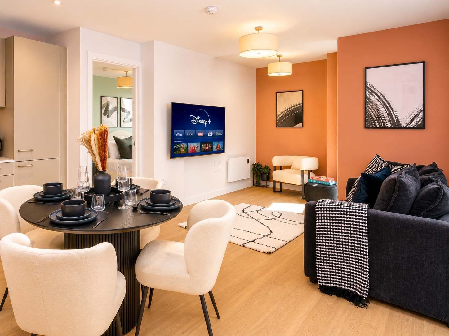 A modern living room with a dining area, a flat-screen TV mounted on the wall, and a sofa with patterned throw.