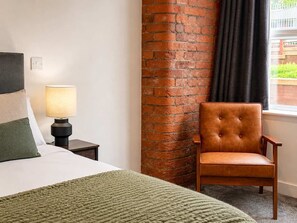 Une chambre d’hôtel avec un lit, une table de chevet avec une lampe, un fauteuil en cuir, un mur en briques et une fenêtre avec des rideaux.