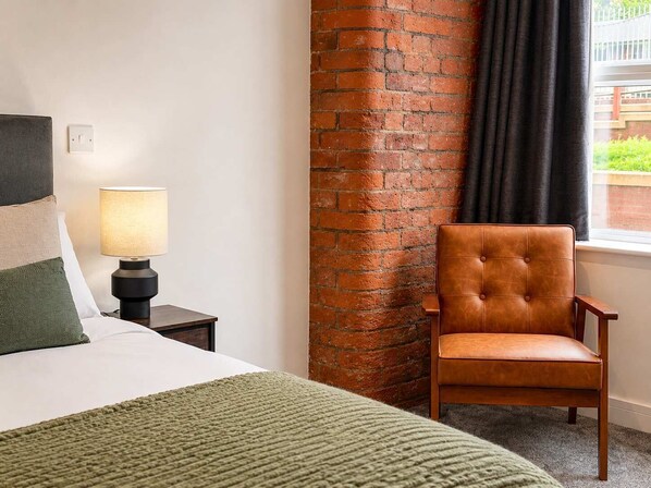 Ein Hotelzimmer mit einem Bett, einem Nachttisch mit Lampe, einem Ledersessel, einer Ziegelwand und einem Fenster mit Vorhängen.