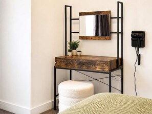 Ein Zimmer mit einem Holztisch, einem Spiegel, einer kleinen Pflanze, einem weißen Hocker und einem an der Wand befestigten Telefon.