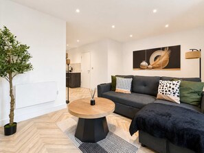 Ein modernes Wohnzimmer mit einem runden Holztisch, einer grauen Couch mit gemusterten Kissen, einem großen abstrakten Gemälde an der Wand und einer Topfpflanze.