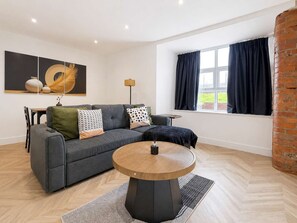 Ein modernes Wohnzimmer mit grauem Sofa, rundem Couchtisch und einem großen Fenster mit Vorhängen.