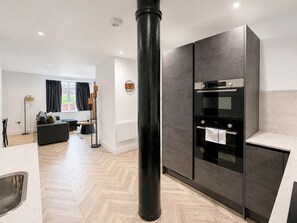 Cuina moderna amb un pilar negre central, forn integrat i zona de menjador amb sofà i làmpada de peu.