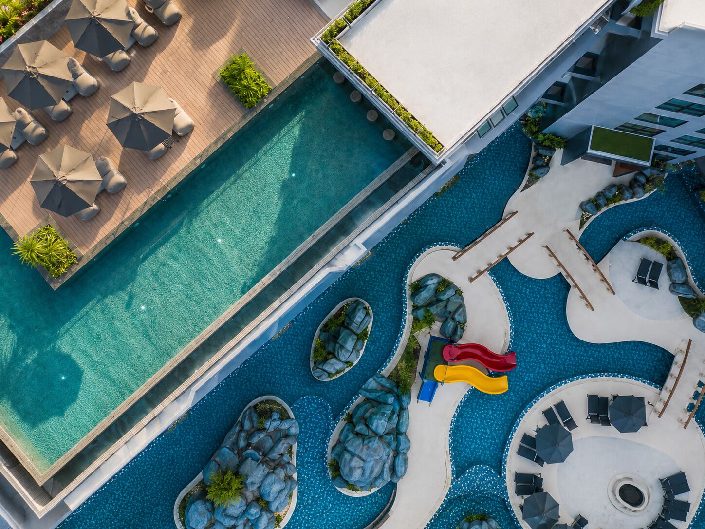 An aerial view of a modern hotel with a swimming pool, sun loungers, and a children's play area.