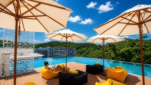 Un área de piscina en la azotea con sillones de descanso amarillos y sombrillas, rodeada de exuberante vegetación y un cielo azul despejado.