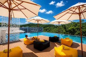Un área de piscina en la azotea con sillones de descanso amarillos y sombrillas, rodeada de exuberante vegetación y un cielo azul despejado.