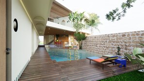 A modern outdoor swimming pool area with a wooden deck, a stone wall, and a bar area visible through an open space. - Hatha House (Tabanan)