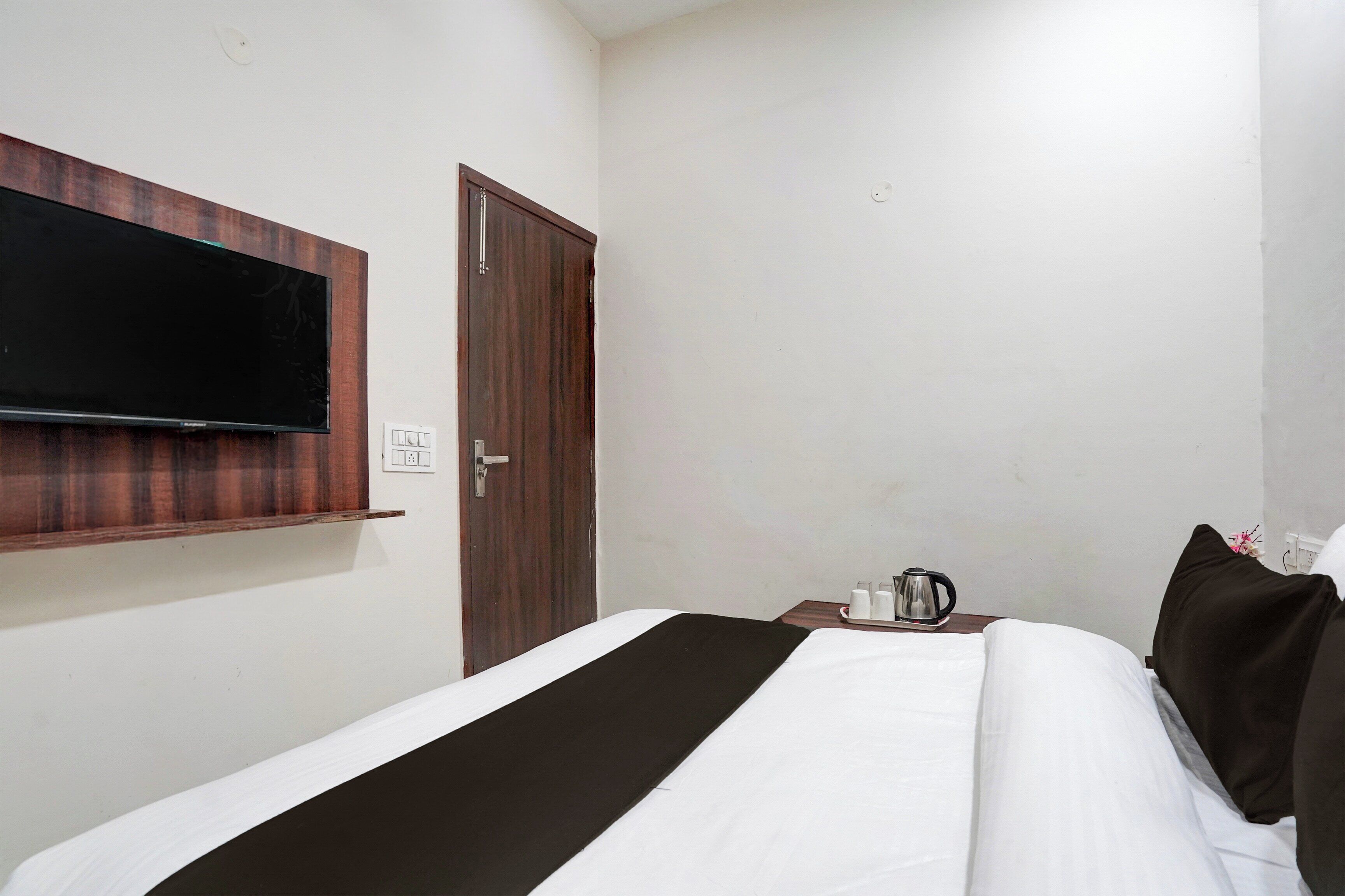 A hotel room with a bed, a flat-screen TV mounted on the wall, a wooden door, and a bedside table with a kettle and cups.
