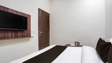 A hotel room with a bed, a flat-screen TV mounted on the wall, a wooden door, and a bedside table with a kettle and cups.
