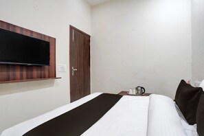 Quarto de hotel com cama, televisão de tela plana na parede, porta de madeira e mesa de cabeceira com chaleira e xícaras.