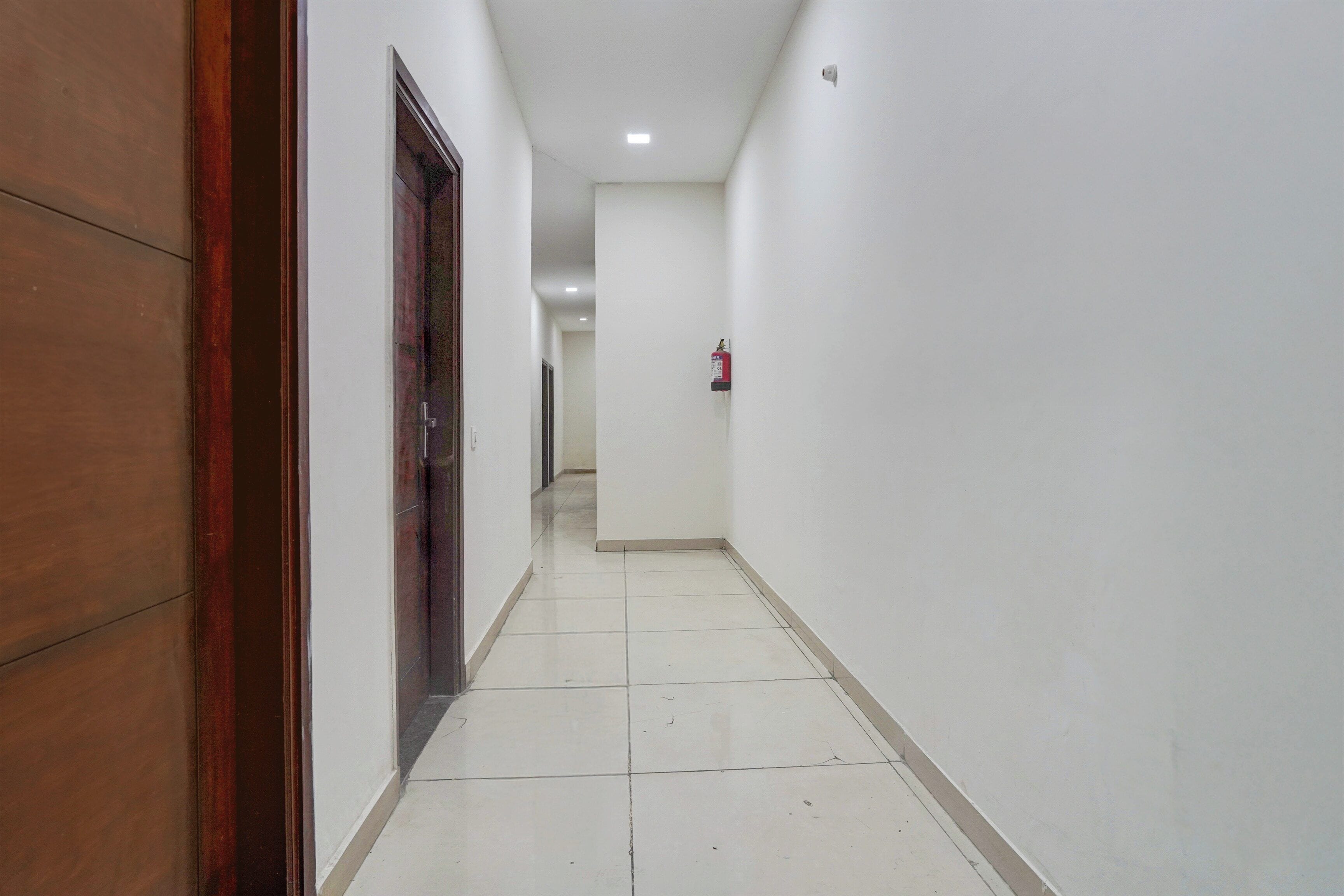 A corridor with white walls, a wooden door, and a fire extinguisher mounted on the wall.