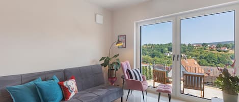 A modern living room with a grey sofa, teal and red cushions, a pink chair, and a large window offering a view of a green landscape and houses.