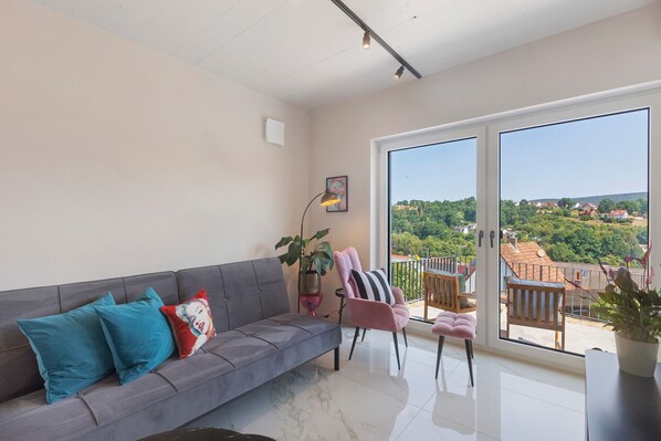 A modern living room with a grey sofa, teal and red cushions, a pink chair, and a large window offering a view of a green landscape and houses.