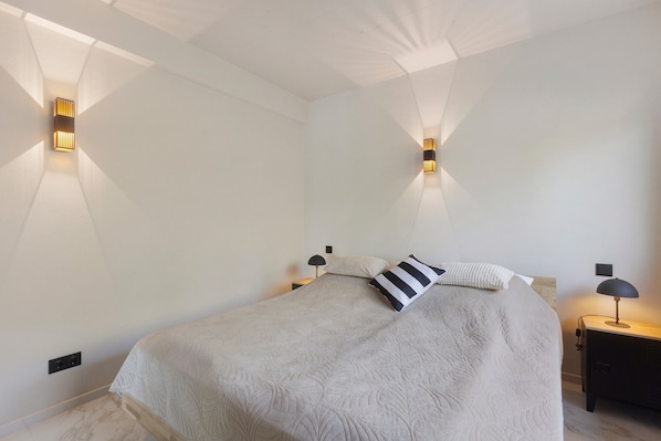 A bedroom with a bed, two bedside tables, a nightstand, and wall-mounted lighting.