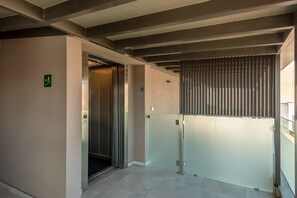 Un ascenseur doté d’une porte en verre, d’un panneau indicateur de sortie de secours fixé au mur et d’une cloison métallique à lattes verticales.